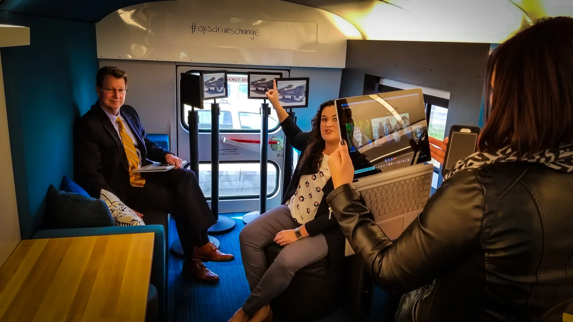 Inside the bus under the #opsdriveschange sign: Superintendent Mark Evans seated on the bench while a video call runs on a laptop.